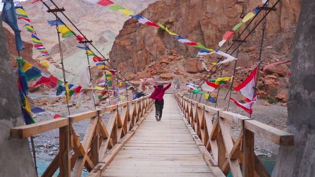 Girl holding shawl and running on a bridge over a clear blue Tsarap river surrounded by mountains in Zanskar, Ladakh, India. Tourist enjoying vacation. Happy people running with joy. Travel concept