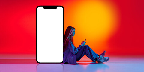Portrait of young charming girl, student sitting near huge smartphone with empty screen over red background in neon light. Concept of online education