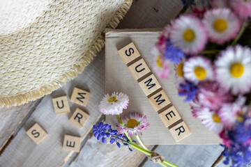 Daisy flowers in white cup composition with a straw hat on wooden background. Summertime, vacations, happiness concept. Flat lay, top view. Letters with word summer on a old book