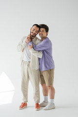 Full length of smiling gay man in casual sweatshirt and shorts hugging bearded boyfriend with...