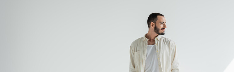 Smiling and young bearded gay man in casual clothes and trendy pearl necklace looking away while...