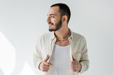 Smiling and bearded gay man in casual clothes and pearl necklace touching shirt and looking away while posing on grey background with sunlight