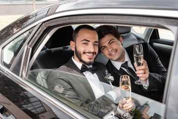 Smiling gay newlyweds in elegant classic attire with boutonnieres holding champagne and looking at...