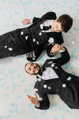 Fototapeta na wymiar Top view of cheerful same sex couple in classic suits holding hands while having fun and lying on confetti during wedding celebration on grey background