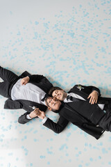Top view of young and smiling homosexual grooms with closed eyes in suits holding hands while lying...