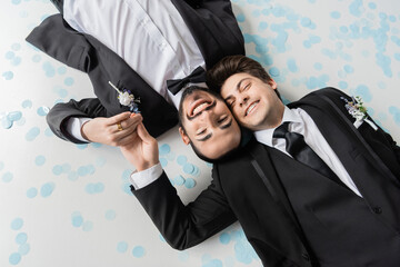 Top view of carefree same sex grooms in elegant suits holding hands and looking at camera while...