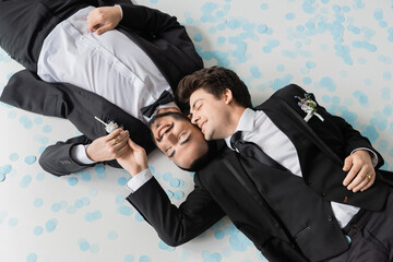 Top view of smiling gay groom touching hand of bearded boyfriend in classic suit lying together on festive confetti during wedding celebration on grey background