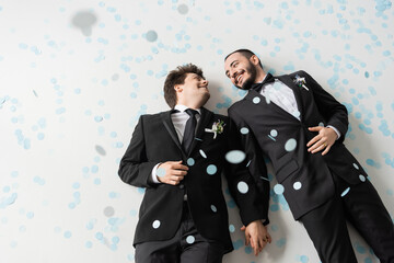 Top view of positive same sex grooms in formal wear looking at each other and holding hands while...