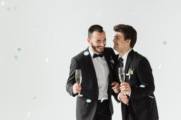 Positive same sex grooms in classic attire holding champagne while standing under falling confetti...
