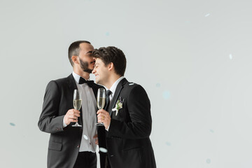 Bearded gay groom in formal wear holding glass of champagne and kissing smiling young boyfriend under confetti during wedding ceremony on grey background