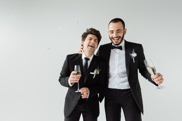 Excited same sex grooms in formal wear with floral boutonnieres holding champagne while celebrating...