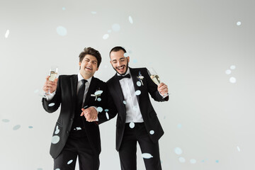 Excited homosexual grooms in elegant formal wear holding hands and glasses of champagne while standing under falling confetti during wedding on grey background