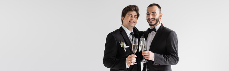 Positive same sex couple in elegant suits holding glasses of champagne and looking at camera during...