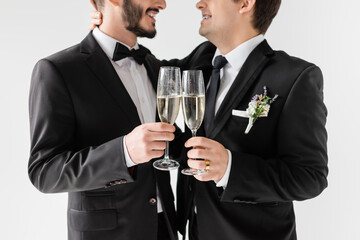 Cropped view of smiling homosexual groom in braces and suit with boutonniere hugging bearded boyfriend and toasting with champagne during wedding ceremony isolated on grey