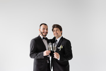 Smiling and young same sex couple in elegant suits with floral boutonniere hugging and holding glasses of champagne during wedding ceremony isolated on grey
