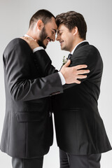 Side view of smiling same sex couple with closed eyes in suits touching each other during wedding celebration while standing isolated on grey
