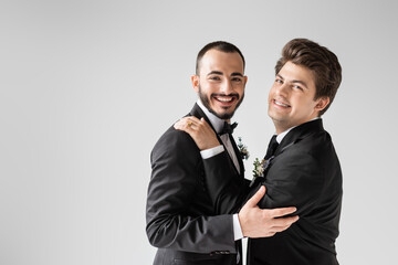 Positive gay man in formal wear with boutonniere hugging young bearded boyfriend in braces and...