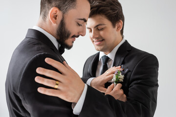 Positive and bearded gay groom adjusting floral boutonniere on elegant suit of blurred boyfriend in braces during wedding ceremony isolated on grey