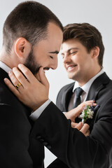 Cheerful bearded gay groom adjusting blurred floral boutonniere on suit of blurred smiling young boyfriend in braces during wedding celebration isolated on grey