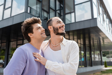 Smiling homosexual man hugging young boyfriend in sweatshirt and braces while looking away together near blurred building  on urban street at daytime