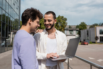 Positive gay man in casual clothes hugging bearded boyfriend using laptop with blank screen near blurred building on urban street at daytime