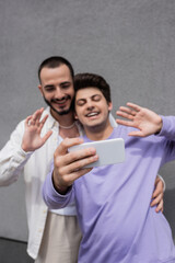 Blurred and young same sex boyfriends in casual clothes having video call on cellphone while standing together near building outdoors