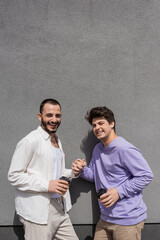 Positive and young same sex couple holding hands and takeaway coffee while looking at camera near grey building outdoors at daytime
