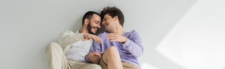 Laughing homosexual couple with closed eyes holding hands and having conversation while sitting together on grey background with sunlight, banner