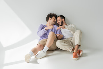 Fototapeta na wymiar Full length of cheerful same sex partners with closed eyes holding hands while talking and sitting together on grey background with sunlight