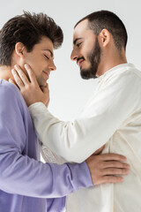Side view of carefree bearded homosexual man in casual clothes touching face of boyfriend with closed eyes while standing together isolated on grey