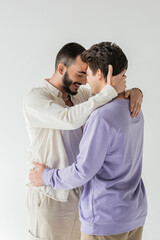 Smiling and bearded gay man in casual beige clothes closing eyes while touching neck and hugging brunette boyfriend isolated on grey