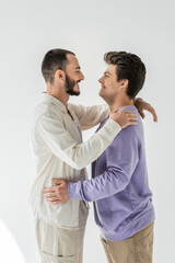 Side view of young and cheerful homosexual couple in casual clothes hugging and looking at each other while standing on grey background