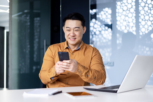 Young handsome Asian man, office worker sitting in the office at the table, fiddling with the phone, typing messages, chatting