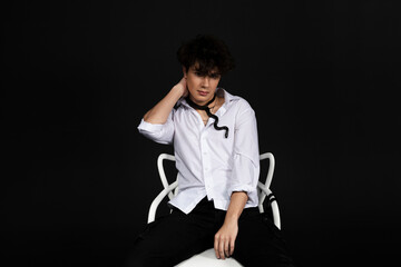 Young handsome man in a white shirt sitting on a chair with a black snake crawling around his neck. Isolated on black background.