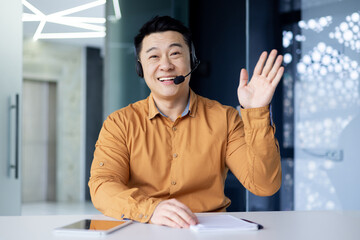 Webcam view, portrait of successful tech support and customer service worker, man smiling and looking at camera waving greeting hello, asian working inside modern office building.