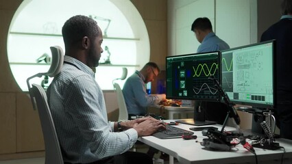 Side view of African American engineer working on innovation project in laboratory. Busy man sitting on work chair and typing code or formula. On screen visible holograms and circuits. Collaboration. - Powered by Adobe