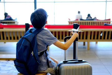 child in airport waiting room, boy of 10 years old in blue shirt with backpack and suitcase holds an airplane, plastic toy in his hands, plays with it, concept of vacation, long journey, air travel
