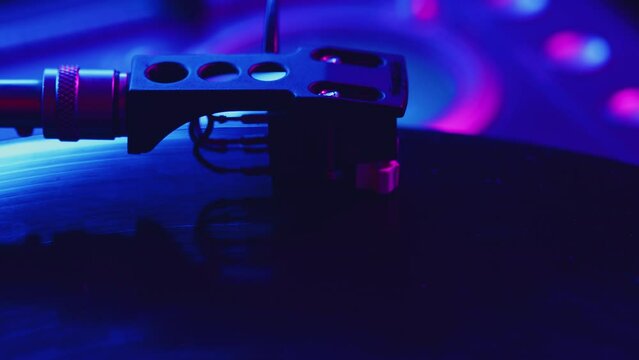 DJ carefully places the needle of the headshell onto the surface of a vinyl record on a turntable and as the needle makes contact, the record begins to rotate close up in a color light