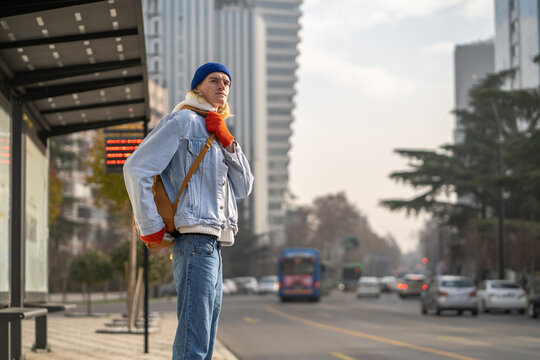 Caucasian guy student with backpack waiting for bus outdoors, young millennial man standing alone at public transport stop, living in big city. Transportation and new millennial generation, city life - Powered by Adobe