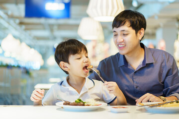 Happy cheerful Asian man and lovely little son enjoy eating a food in the fast food restaurant together. Man looking his son eating a meat ball.