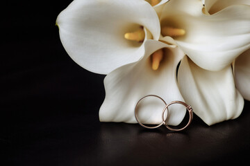 Minimalistic wedding bouquet of white zontedexia or calla flowers. The bouquet lies on a dark surface on a black background with gold rings with two gold rings.