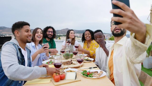 Happy young group of multiracial friends enjoying barbecue dinner party at house terrace. Millennial cheerful men and women taking selfie portrait with phone during birthday celebration.