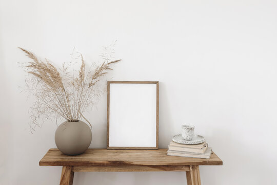 Ball shaped vase with dry grass bouquet. Old wooden bench. Blank vertical picture frame mockup with books and cup of coffee, tea. White wall background. Empty copy space. Neutral still life interior.