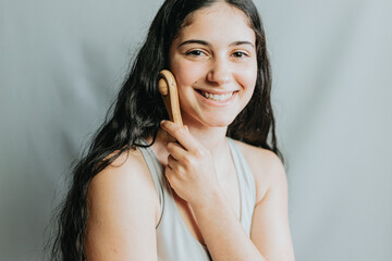 Young woman applying facial massage with wooden roll. Joyful selfcare day. Applying pressure to improve circulation. New methods of skincare and daily rituals.Portrait in studio with grey background.