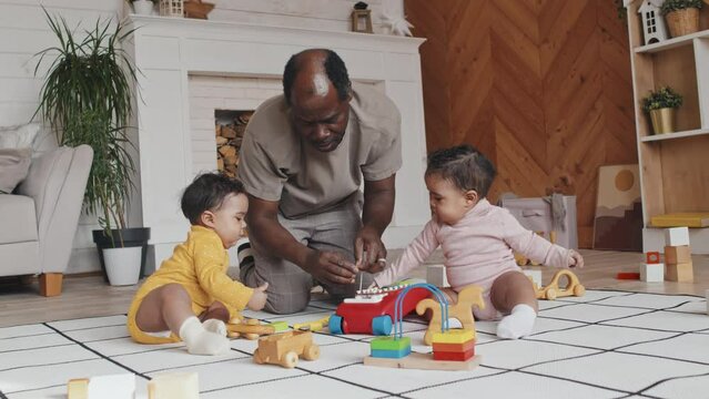 Two little babies and African American father playing with toys on floor in nursery room