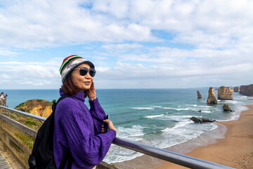 Asian woman in hat, sunglasses, backpacking, standing at scenic seaside viewpoint in Port Campbell National Park. Great Ocean Road, Victoria State, Australia.