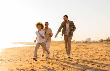 Beach fun. Happy diverse family walking and fooling by the seaside, boy running along beach being...