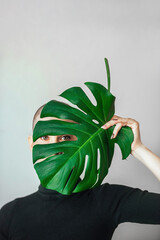 Portrait of an emotional young woman with a shaved bald head and a large green leaf of a flower behind which she hides. Result of chemotherapy during cancer treatment