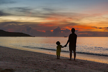 Fototapeta na wymiar Mother and her little child walking down the beach park holding hands at sunset