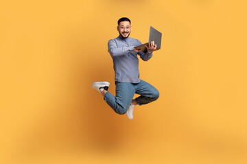 Happy asian man with laptop computer jumping up over yellow background